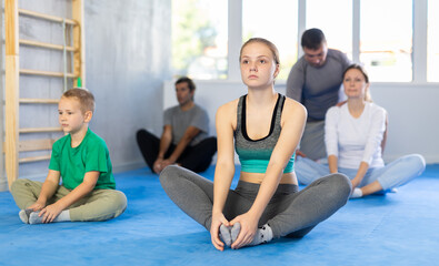 Girl with her family sitting on mat in butterfly pose, stretching inner part of thigh during group...