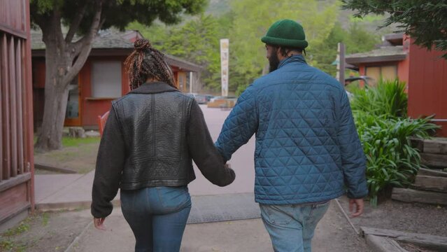 Back view of a diverse African American couple holding hands and walking down a peaceful street, with rustic cabins and lush greenery around. Slow Motion, Camera 4K RAW. 