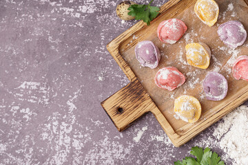 Fototapeta na wymiar Wooden board with colorful raw dumplings and flour on dark background