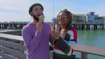 A black man in a green beanie and black woman with braided hair laugh while eating ice cream on a sunny pier overlooking the sea. Slow Motion, Camera 4K RAW. 