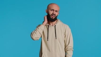 Sick middle eastern guy suffers from neck pain on camera, dealing with sore throat and muscle pressure over blue background. Adult being unwell with spine discomfort in studio. Camera 1.