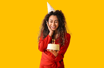 Beautiful young African-American woman in party hat with sweet cake celebrating Birthday on yellow background