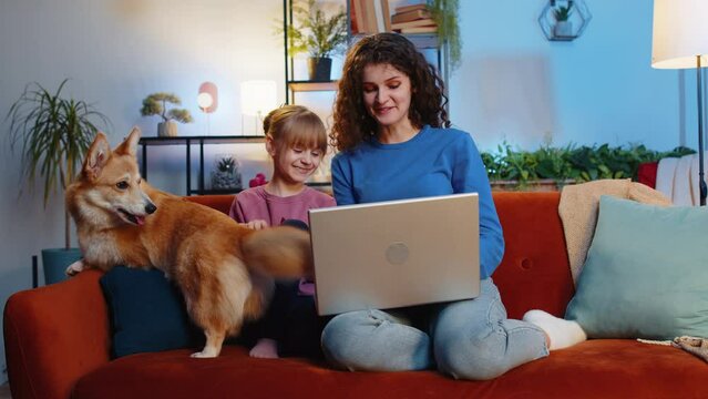 Smiling mother and daughter child girl using laptop sitting beside cute corgi dog on sofa in living room. Family watching movie together on netbook during weekend at home. Online education concept.
