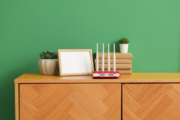 Modern wi-fi router with books, blank frame and plants on commode near green wall
