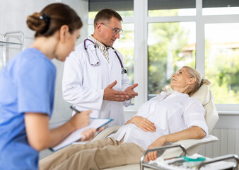 Obraz premium Male doctor and female assistant having conversation with elderly woman patient about need for additional diagnostic procedures, long-term and comprehensive treatment, assistant records data in report