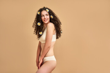 Self care. Sensual caucasian lady dressed in only underwear looking aside while posing over beige background. Young skinny woman wearing chamomile bloom in curly dark hair in studio. Copy space. - obrazy, fototapety, plakaty