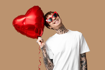 Handsome young man in sunglasses with heart-shaped air balloon on beige background