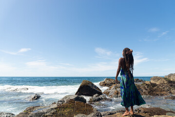 Beautiful woman, with braids and blue clothes, standing on the rocks at the beach posing for a photo