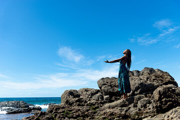 Beautiful woman, in blue clothes and braided hair, standing on the dark rocks of the beach with open arms
