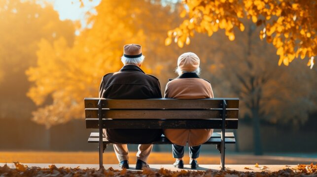 An elderly couple sits together on a park bench, surrounded by golden autumn leaves, in a serene, peaceful setting.