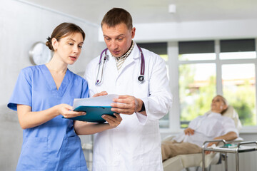 Man doctor gives instructions to girl nurse about diagnosis of patient, medical specialists consider outpatient card, anamnesis questionnaire. Blurry client in background - obrazy, fototapety, plakaty