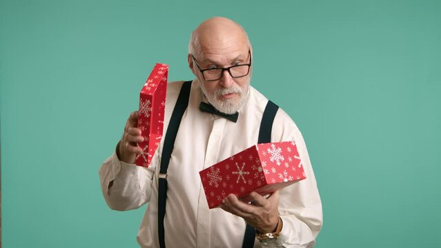 Cheerful older man with a white beard and suspenders opens a red gift box, his face expressing childlike joy on a teal background. High quality 4k footage