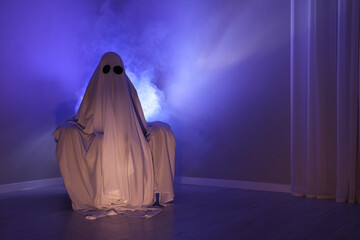 Creepy ghost. Woman covered with sheet sitting in armchair in blue light, space for text