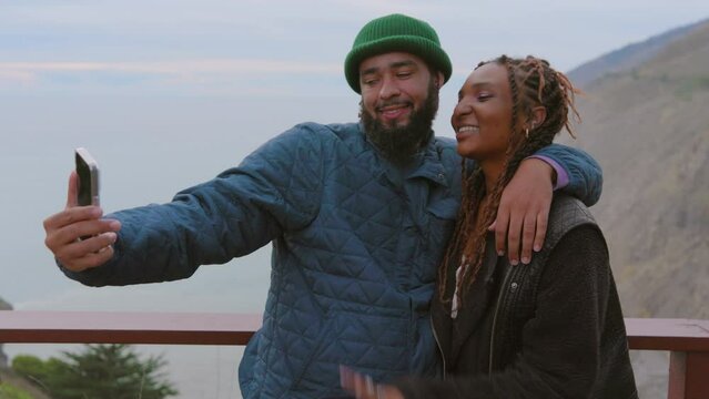 Black couple in warm clothes shares a selfie moment on a scenic overlook, with a vast ocean and cloudy sky in the backdrop. Slow Motion, Camera 4K RAW. 