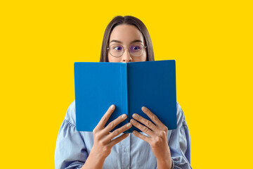 Young woman in eyeglasses reading book on yellow background