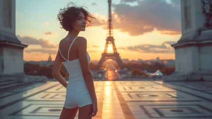 Rolgordijnen European woman model short hair and smiling face wearing a fitted casual white color short slip dress with on the background the Eifel tower of Paris at sunset © Chirapriya