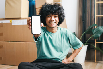 Cheerful young Asian man sitting crossed legs on the floor and showing white blank phone screen...