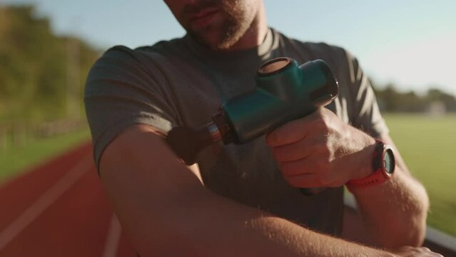 Athlete massages with percussion therapy gun to relieve pain of muscle aches after workout. Man massages his body with massage percussion device after training session. Muscle warming up with massager