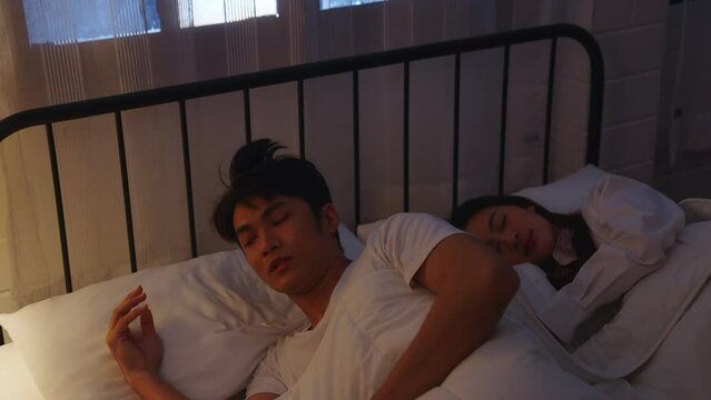 Asian couple sleeping in bed. Cheating husband. Couple issue. Infidelity betrayal. Disloyal man using mobile phone for chatting to lover lying in bed with sleeping wife late at night in warm light.