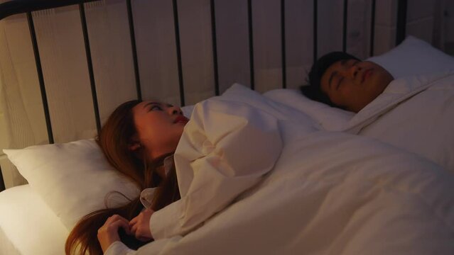 Asian couple sleeping in bed. Cheating wife. Couple issue. Infidelity betrayal. Disloyal woman using mobile phone for chatting to lover  lying in bed with sleeping husband late at night in warm light.
