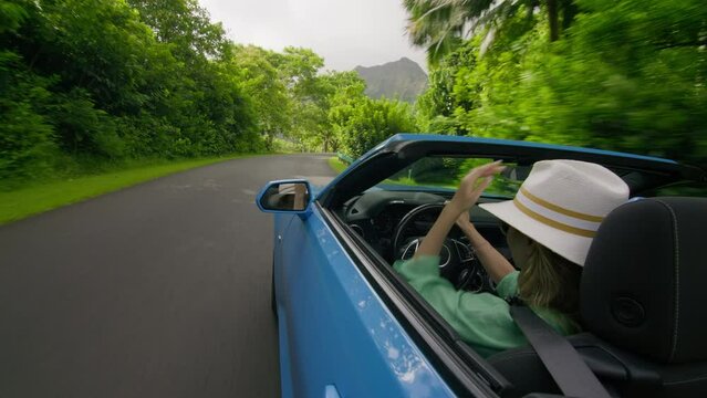 View from outside blue convertible car on stylish young woman driver. Back view happy traveler female waving hand, catching air while driving fast by scenic green tropical island. Vacation background