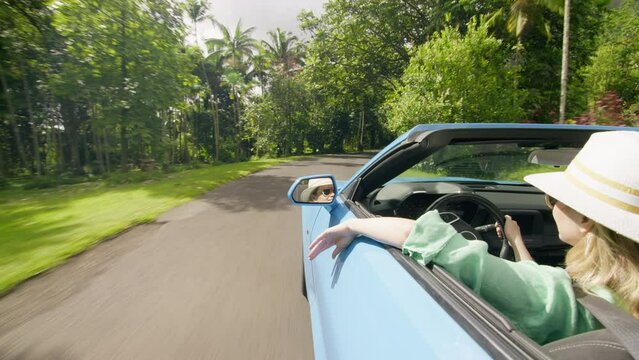 Vacation on scenic green tropical island background. View from outside blue convertible car on stylish young woman driver. Back view happy traveler female waving hand, catching air while driving fast