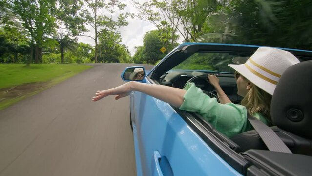 Back view happy traveler female waving hand, catching air while driving fast by scenic green tropical island. Vacation background. View from outside blue convertible car on stylish young woman driver