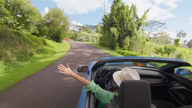 Free girl with hand out of convertible car rides with wind blowing in face. Stylish tourist stretches hand out of rent car without roof. Woman driving in botanical tropical garden on Hawaii road trip
