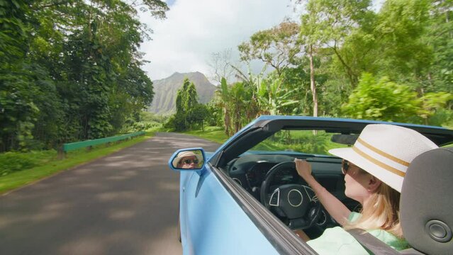 Happy girl in blue convertible car enjoying summer day and catching wind with hand. Stylish woman travels by tropical island with mountain landscape. Hawaiian tourist stretching hand out to sun rays