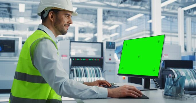 Caucasian Male Industrial Engineer In Protective Gear Working on a Desktop Computer with Green Screen Chromakey on Display at an Electronics Factory. Technological Research and Development Center.