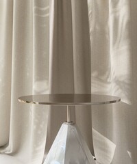 Close up of shiny, modern round gold podium table, marble leg with beige drapery curtain in...