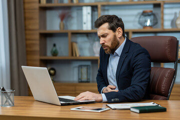 Obraz premium Unhealthy businessperson holding hand on stomach and grimacing in pain while sitting in workspace interior. Handsome male feeling unwell and having severe belly ache due to skipping full meals.