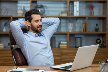 Relaxed brunette man leaning back and holding hands behind head in personal workspace with pc. Calm...