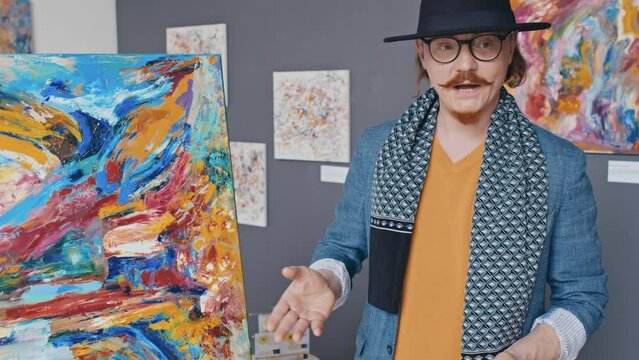 Medium tilt-up shot of eccentric young Caucasian male artist with red beard, moustache, in fedora hat, scarf, glasses demonstrating new abstract artwork and describing its meaning at public event