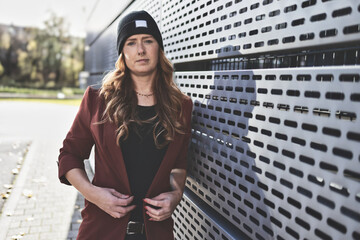 Woman in her mid-thirties with long red hair wearing black clothing with a brown jacket and hat in...