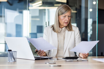 Concentrated and worried scary gray-haired businesswoman sitting in the office at the desk, reviewing and holding documents in her hands. Problems with financial and bank accounts.