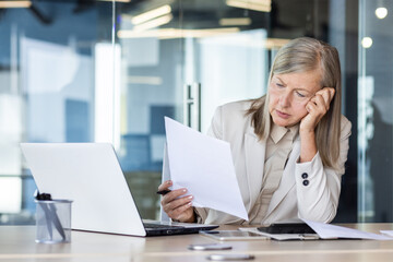 Tired and upset senior woman in suit working in office. He sits at the desk, leans his head on his...