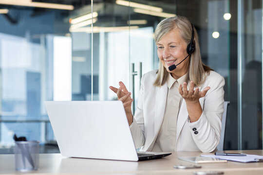 Senior smiling gray-haired woman in a suit and headset works in a call center, sits in the office at a laptop, consults and conducts an online video meeting, smiles and gestures with her hands.