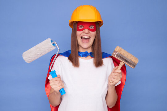 Skilled painter. Home improvement. Interior decorator. Excited woman painter wearing superhero costume and protective helmet holding painting roller and brush isolated over blue background