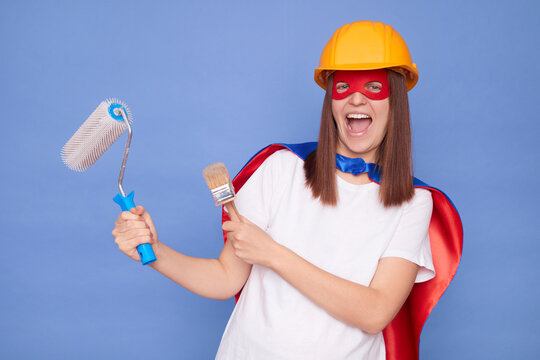 Excited woman painter wearing superhero costume and protective helmet holding painting roller and brush enjoying her painting work during house improvement isolated over blue background