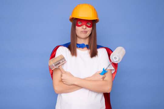 Exhausted sad woman painter wearing superhero costume and protective helmet holding painting roller and brush isolated over blue background being tire from house decoration work