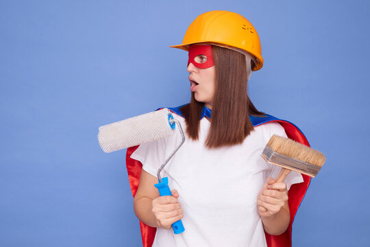 Shocked woman painter wearing superhero costume and protective helmet holding painting roller and brush isolated over blue background looking aside with surprised face