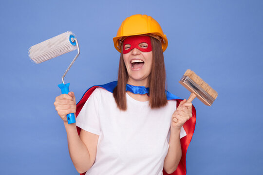 Professional painter. Decorative painting. Building service. Overjoyed woman painter wearing superhero costume and protective helmet holding painting roller and brush isolated over blue background