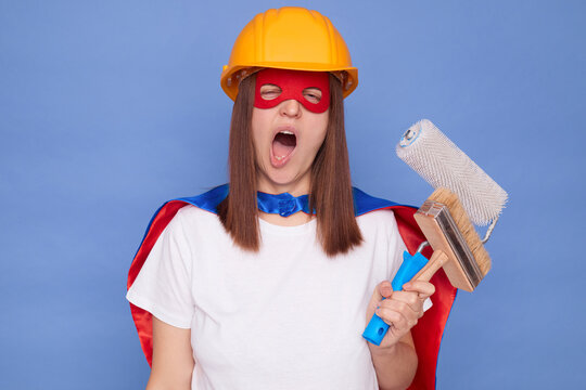 Sleepy overworked woman painter wearing superhero costume and protective helmet holding painting roller and brush isolated over blue background yawning after house painting and renovation work