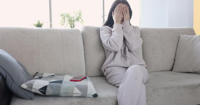 Upset young woman sitting on sofa waiting for message on smartphone. News. Stressed woman thinking about sudden problems or relationship breakup