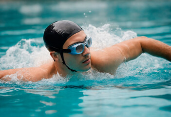 Fototapeta na wymiar Young man in goggles and cap swimming butterfly stroke style in the pool
