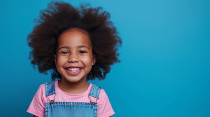 young African American girl with a big smile, wearing a pink shirt and a blue denim jumper, set against a light blue background