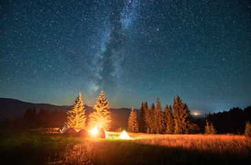Night camping in mountains under starry sky and Milky way. Tents standing in campsite, fire burning, smoke raising to sky, stars shining. Concept of hiking in mountains. - obrazy, fototapety, plakaty