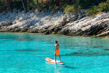 Fit man standing on a SUP board or paddleboard, floating on the clear blue water, looking away.