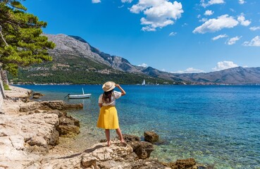 Back view of woman in stylish summer clothes standing on rocky beach in old Korcula town, Croatia